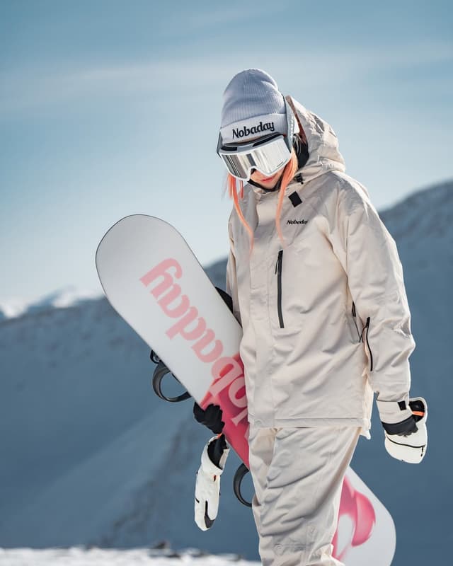A snowboarder with pink hair, wearing a beige suit, white Nobaday beanie and goggles, holds a white Nobaday snowboard in a snowy mountain landscape.