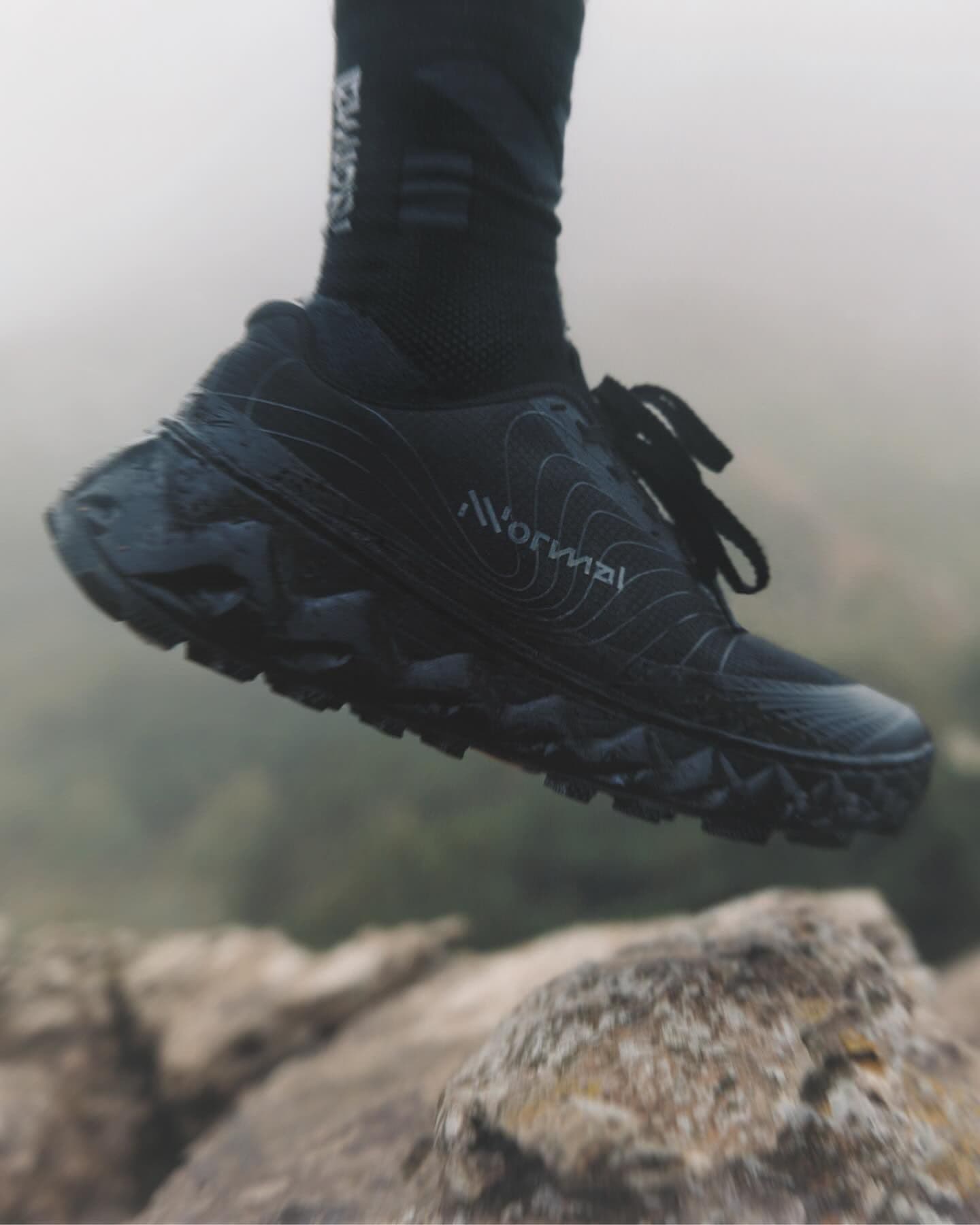 A black athletic shoe with "NORMAL" on its side, mid-air above rocks in a foggy outdoor setting.