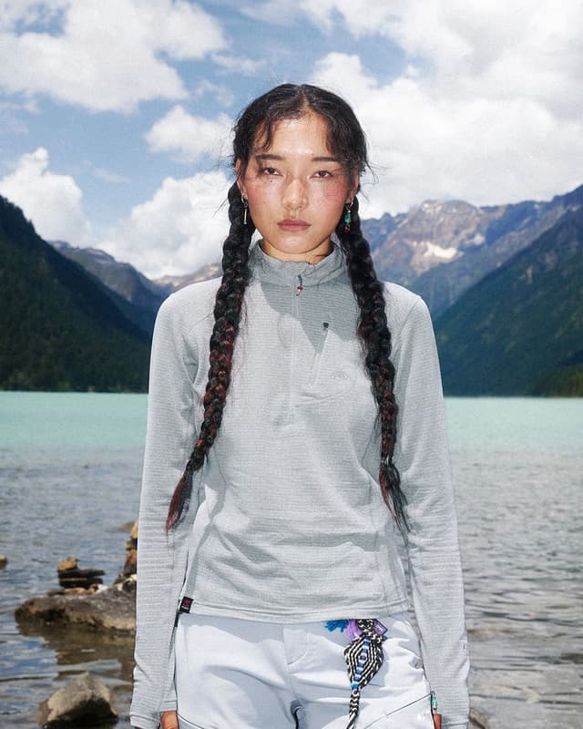 A young woman with two long braids, wearing a gray long-sleeved top, stands in front of a turquoise mountain lake.