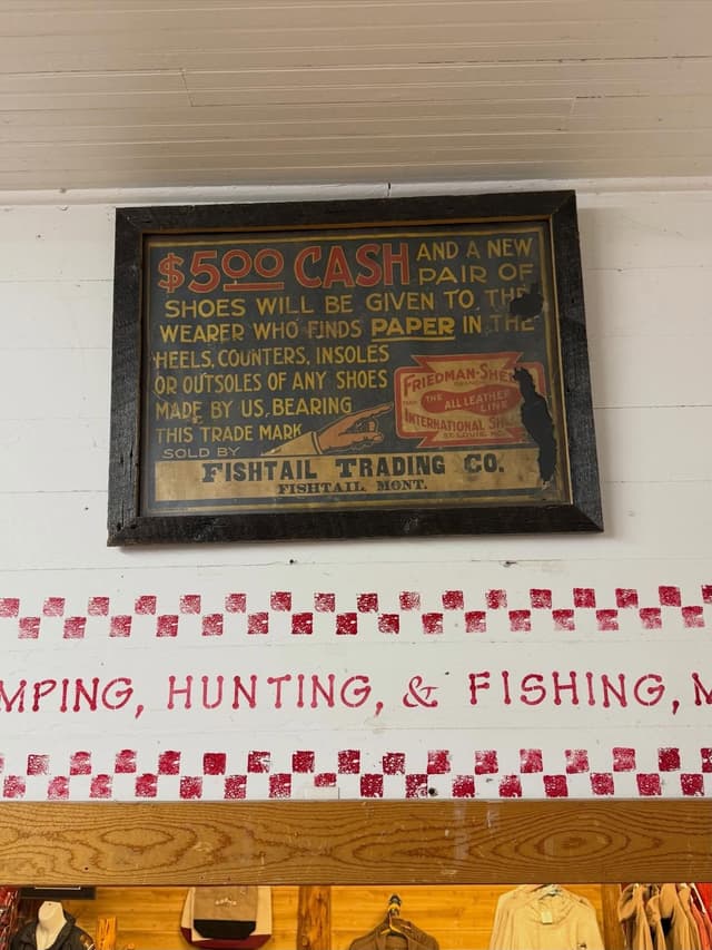 Vintage sign offering "$500 CASH and new shoes" for finding paper in Friedman-Shelby footwear, sold by Fishtail Trading Co. Below, wall decor includes red checkered patterns and "CAMPING, HUNTING, & FISHING."