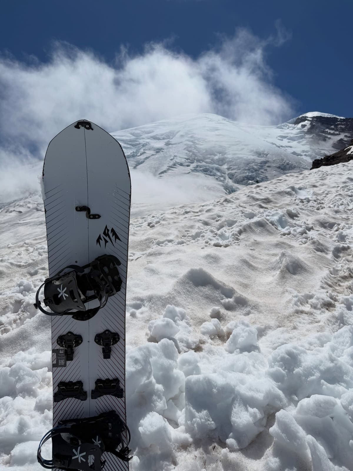 A white splitboard stands in patchy snow with a large, snow-covered mountain and blue sky in the background.