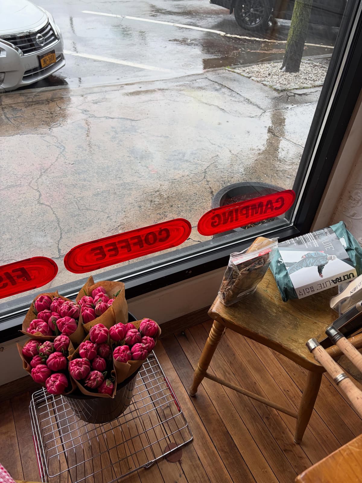 A view from inside a shop on a rainy day, showing a bucket of pink tulips and fishing rods near a window with "COFFEE" and "CAMPING" stickers, overlooking a wet street.