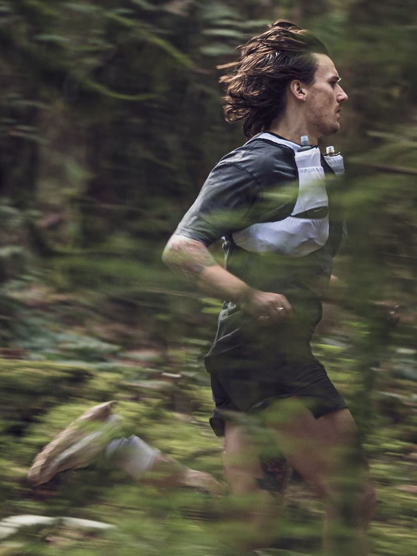 A man trail running through a blurry green forest, wearing a white hydration vest with two bottles.