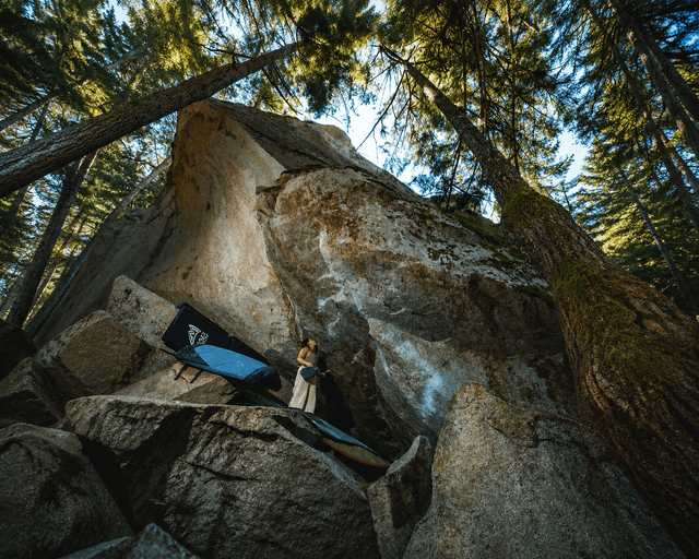 A woman stands on a rock next to bouldering crash pads, surrounded by giant boulders and tall trees in a sunlit forest.