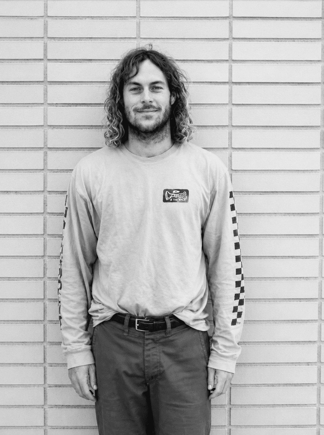 A smiling man with curly hair and a beard, wearing a long-sleeved shirt with checkerboard sleeves, stands against a tiled wall.