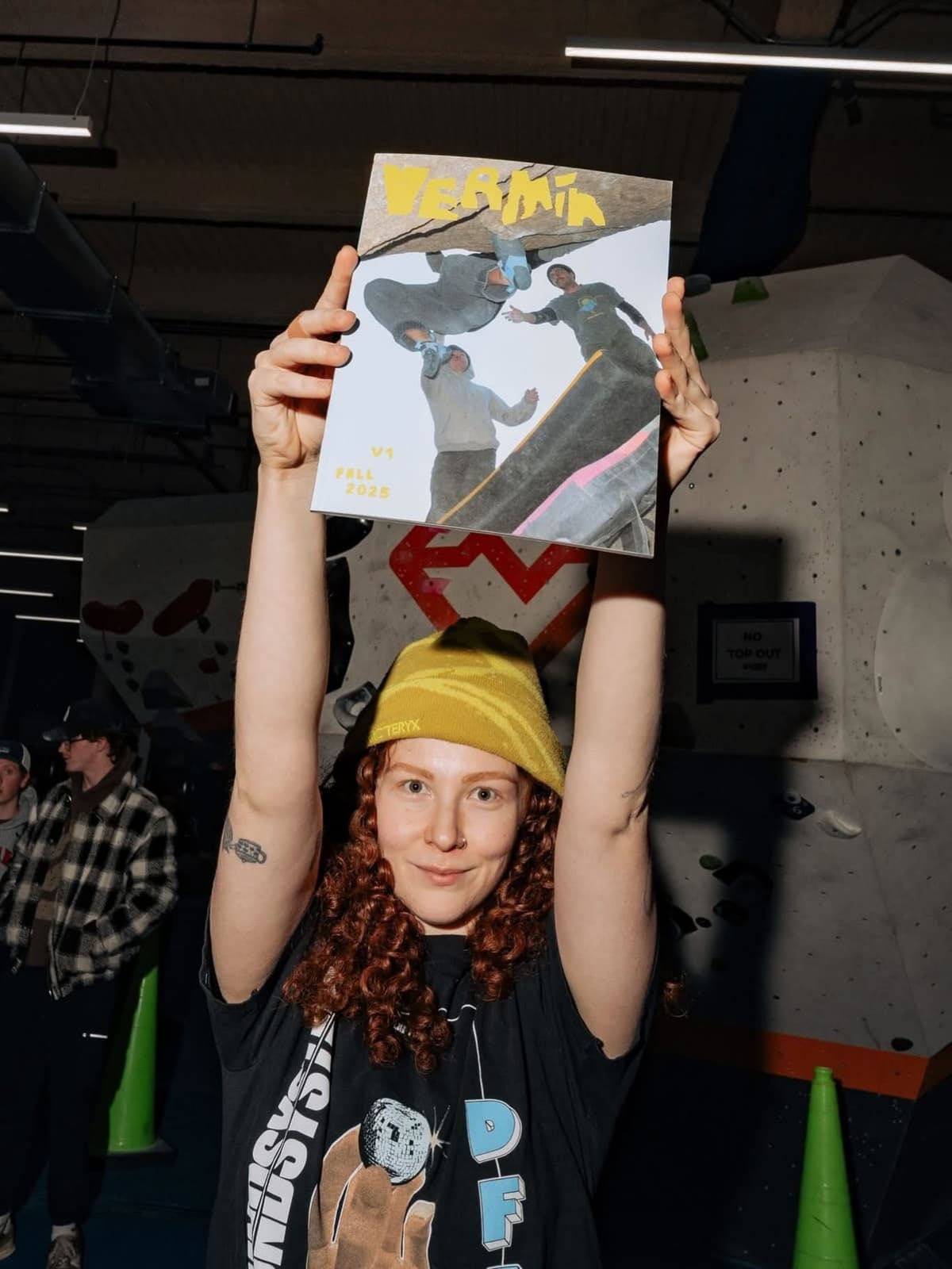 A woman with red curly hair and a yellow beanie holds a "VERTICIA" magazine with climbers on its cover above her head in a climbing gym.