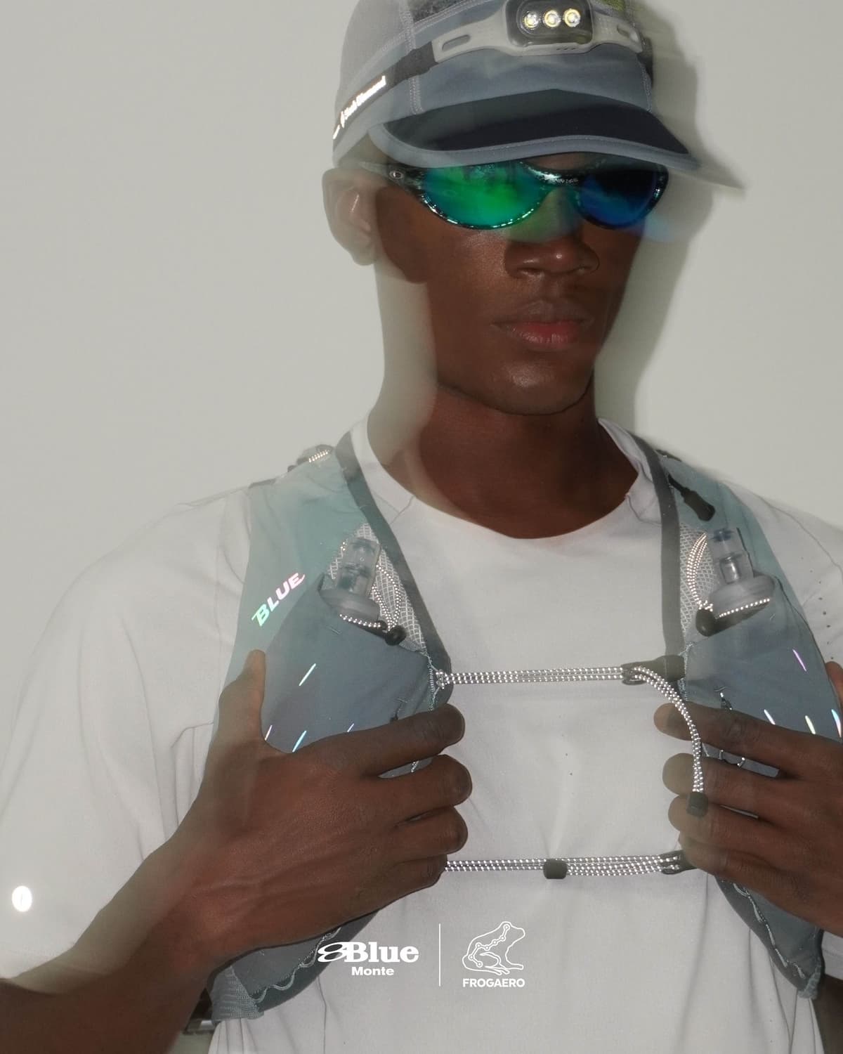A person wearing a cap with a headlamp, green mirrored sunglasses, and a slate blue running vest with hydration bottles.
