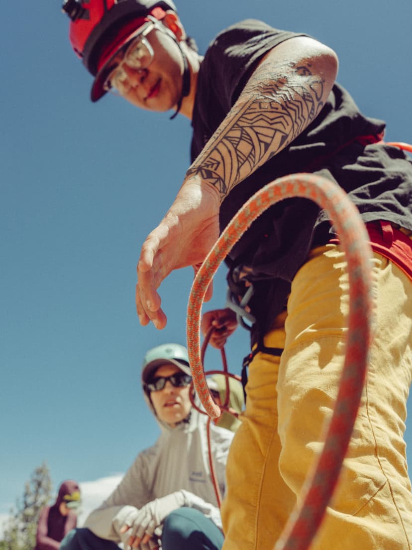 A climber in a red helmet, glasses, and a geometric arm tattoo, wearing yellow pants, holds a rope against a blue sky. Another person is blurred in the background.