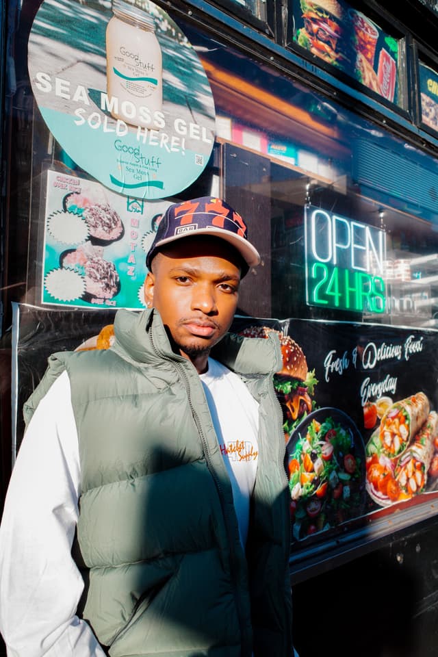 A man wearing a cap and green puffer vest stands in front of a store window with food advertisements and a neon "OPEN 24 HRS" sign.