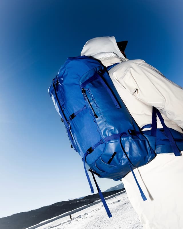 Person in white winter clothing and a blue backpack, viewed from behind against a clear blue sky and snowy landscape.