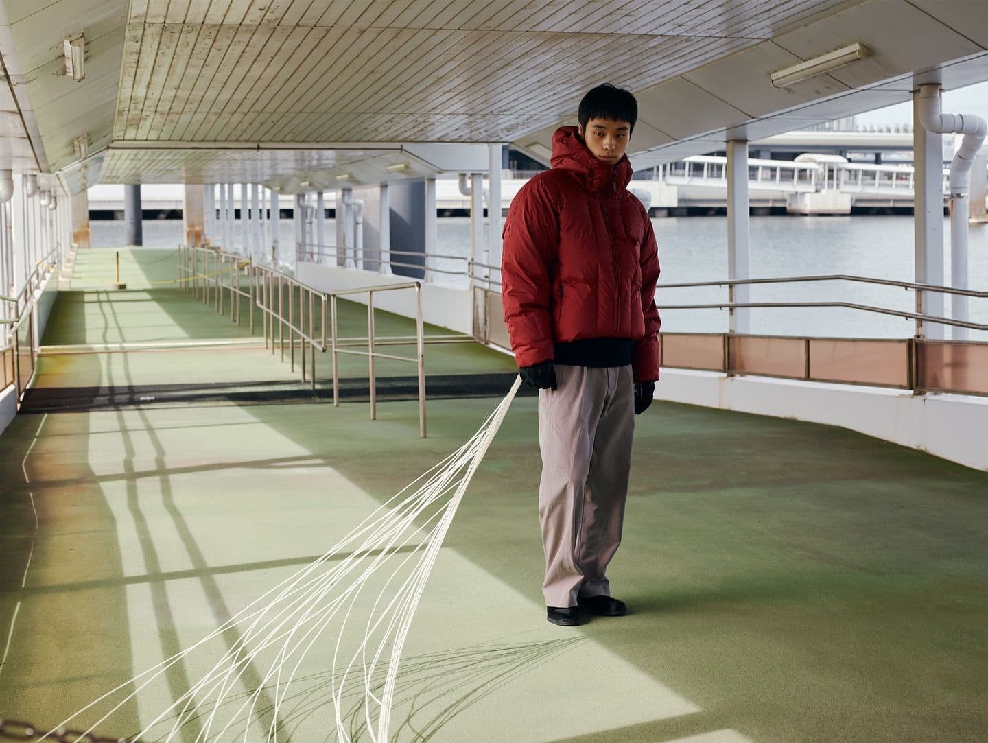 A man in a red puffer jacket stands on a covered pier, holding white strings fanning out on the ground behind him.
