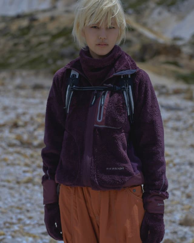 Blonde woman in purple fleece jacket, orange pants, and gloves in a rocky outdoor landscape.