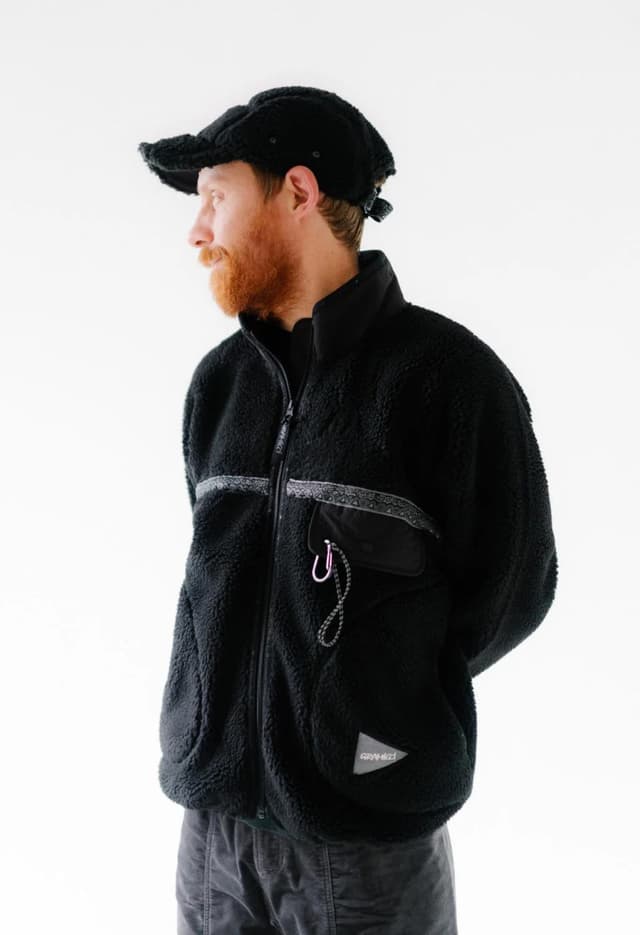 A man with a red beard and black cap wears a black fleece jacket with a chest pocket and pink carabiner, looking left.