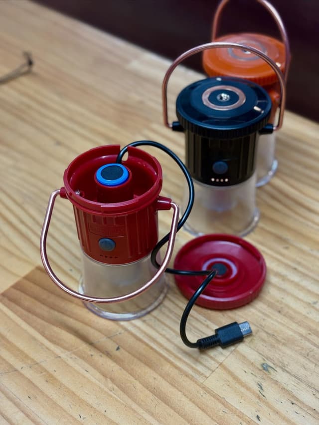 Three red, black, and orange portable lanterns on a wooden surface; the front red lantern is open with a USB cable plugged into its top.