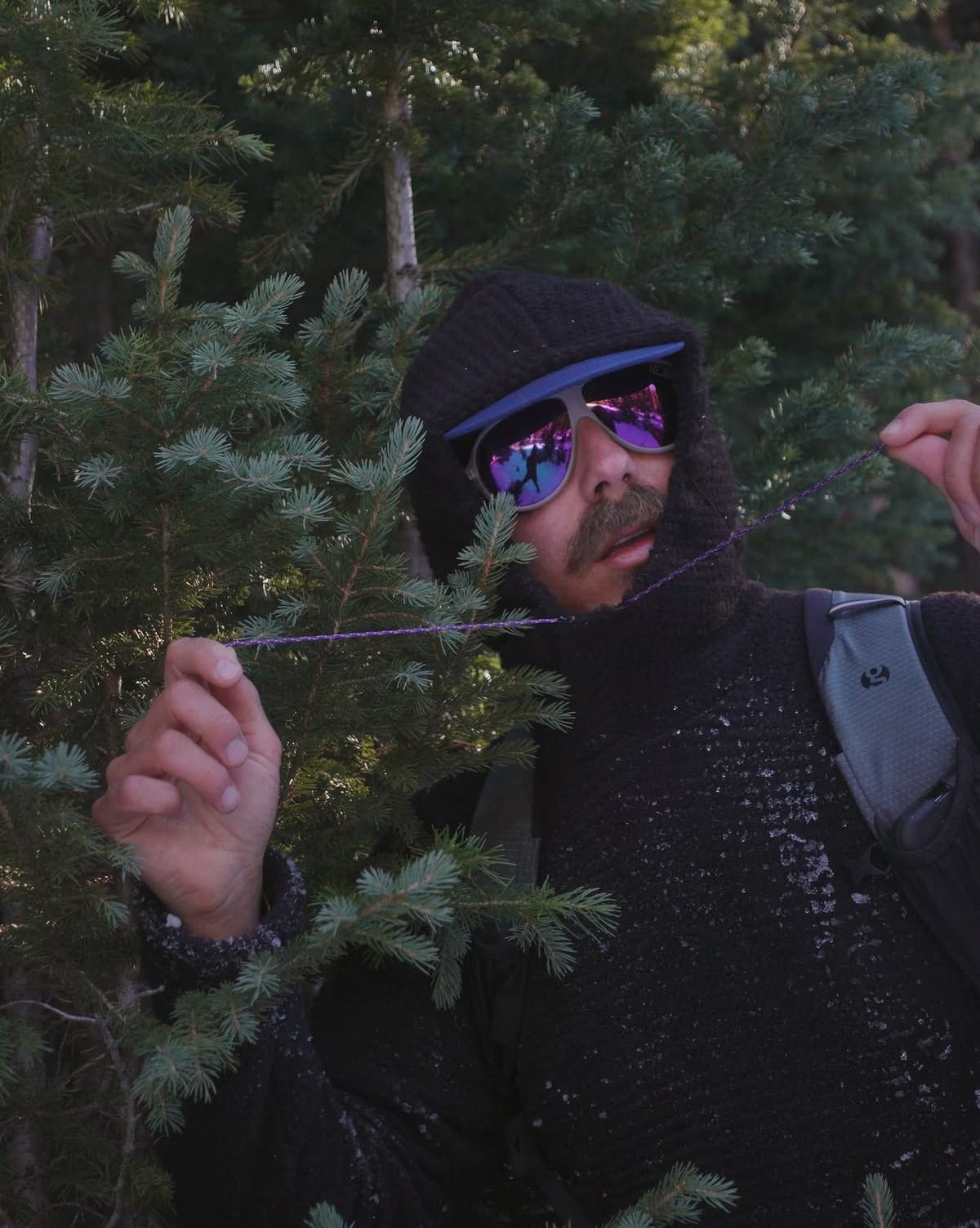 A person in a black knitted hoodie and reflective purple sunglasses stands among pine trees, holding a thin purple string, with frost flecking their clothing.