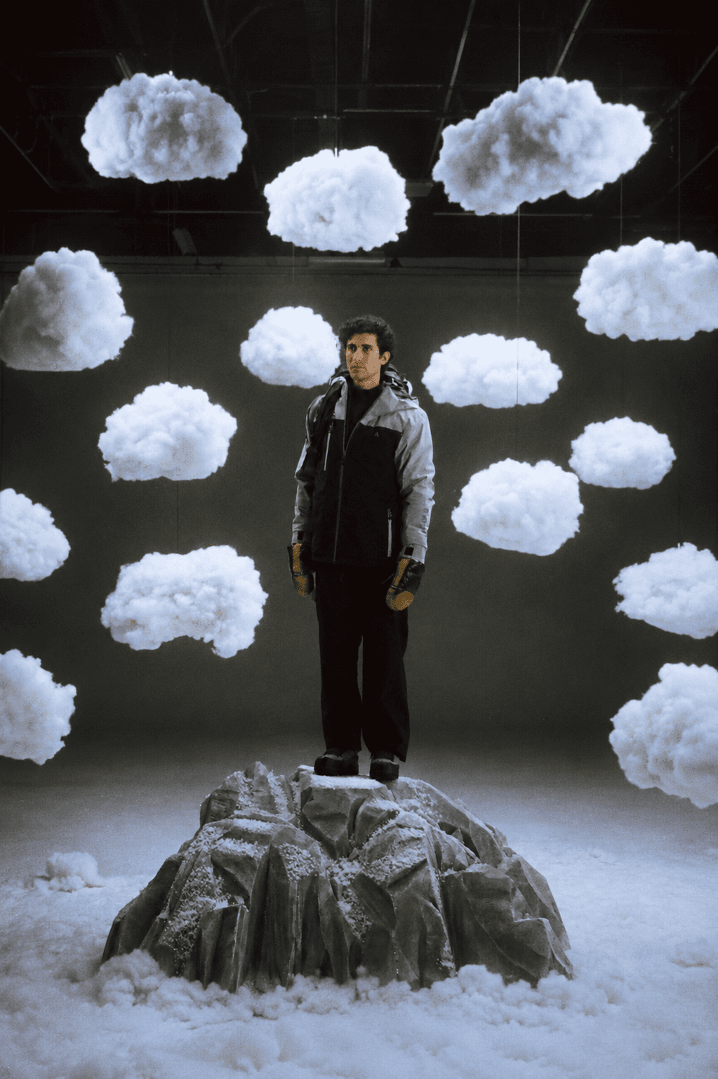 A man in winter gear stands on a snowy rock surrounded by hanging artificial clouds in a studio.