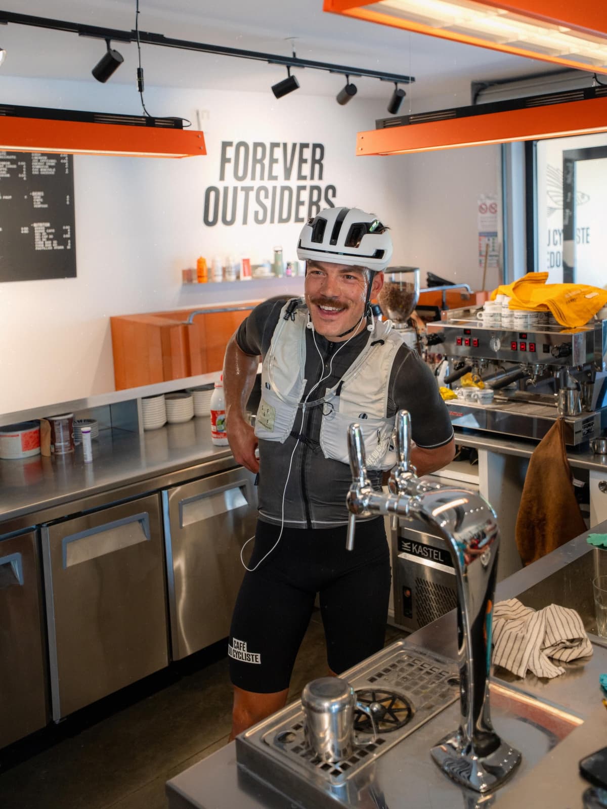 Smiling cyclist in a helmet and vest standing behind a cafe counter with a "FOREVER OUTSIDERS" sign.