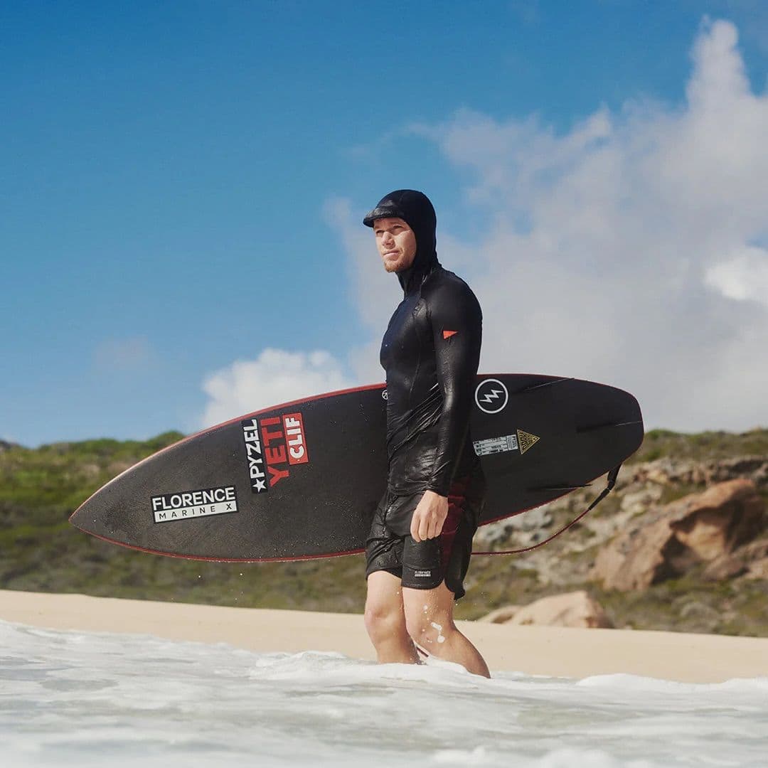 Man in a black hooded wetsuit top and shorts carrying a black surfboard, walking out of ocean waves onto a sandy beach.