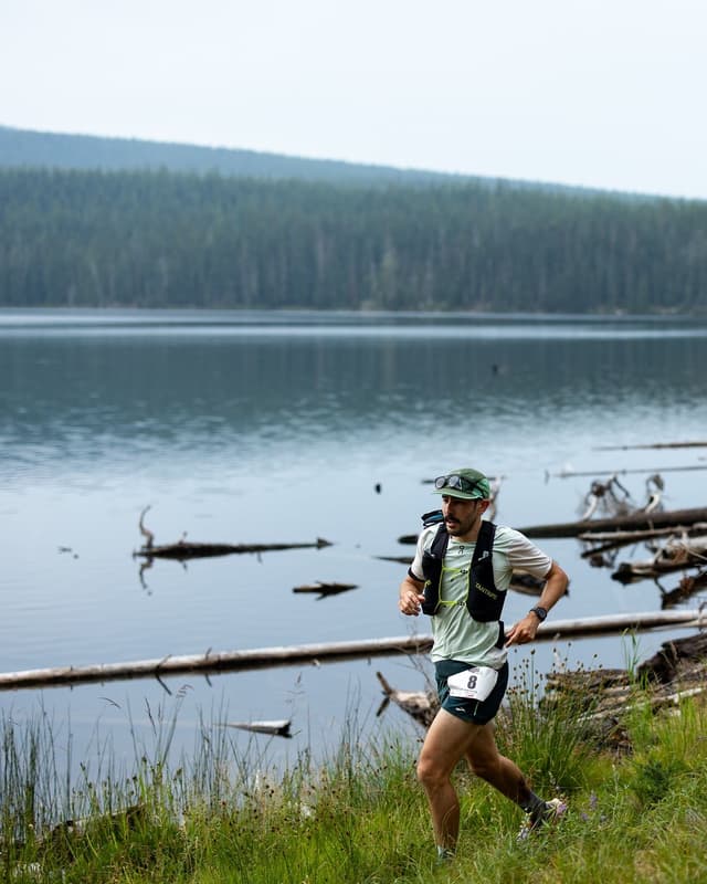 A male runner wearing a hydration vest and race bib runs on a grassy trail beside a lake with a forest in the background.