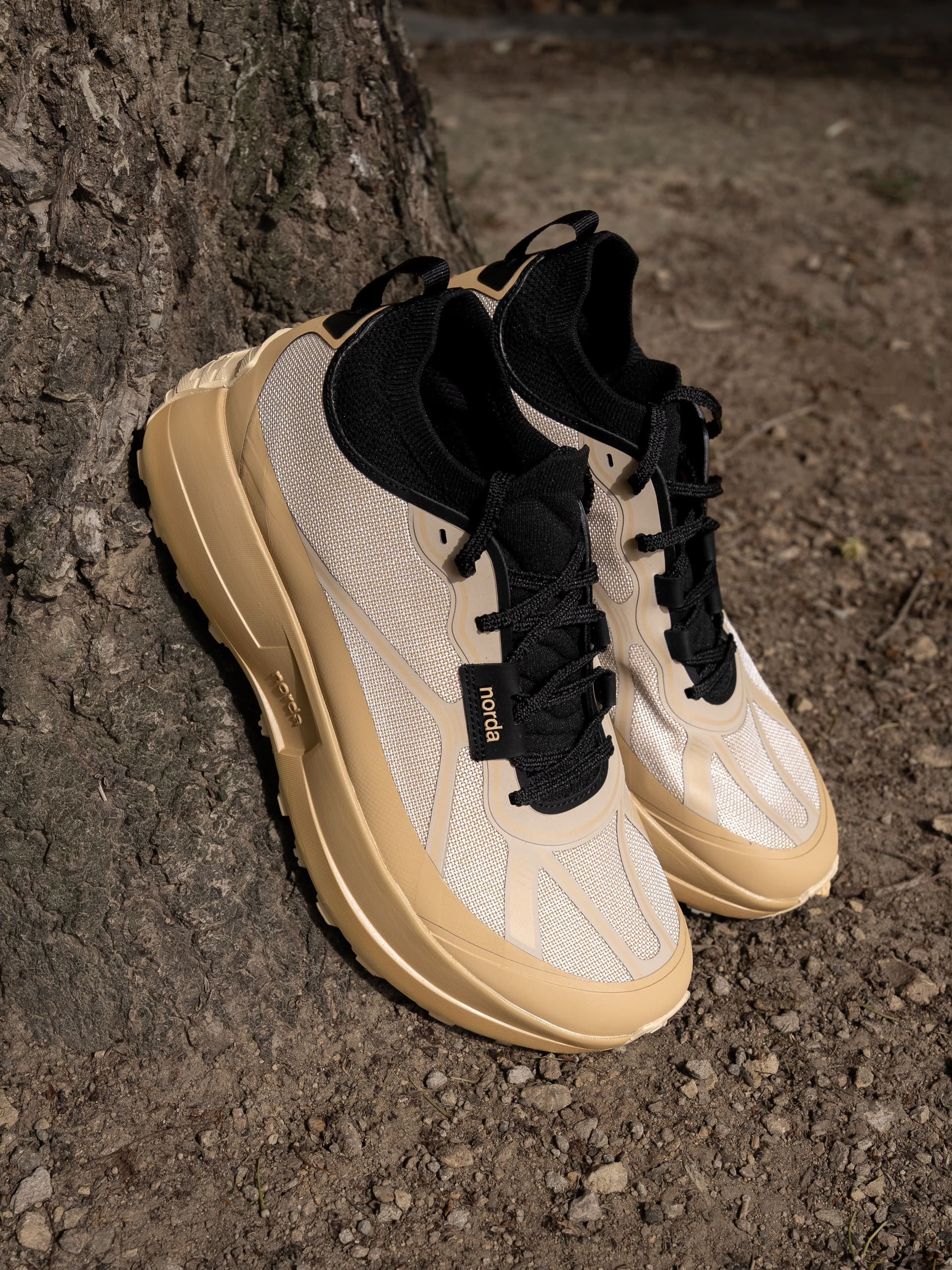 A pair of beige and black Norda trail running shoes leaning against a tree trunk.