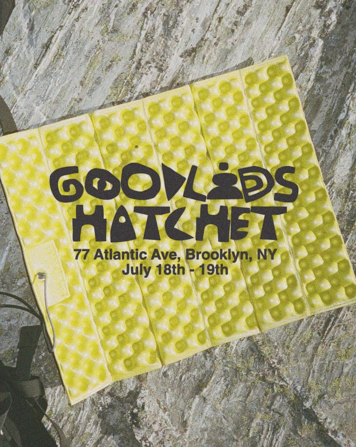 A yellow textured camping pad on a rocky surface featuring text: GOODLED HATCHET, 77 Atlantic Ave, Brooklyn, NY, July 18th - 19th.