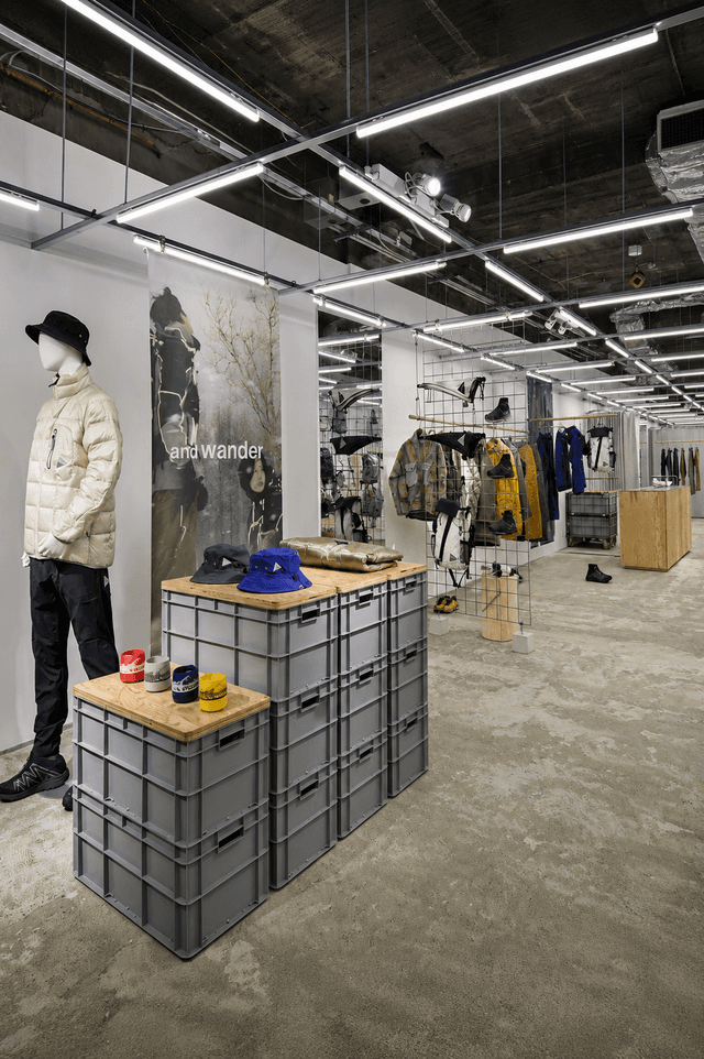 Interior of a minimalist clothing store with concrete floors and ceiling, a mannequin in a puffer jacket, and displays made of stacked grey crates under linear fluorescent lighting.