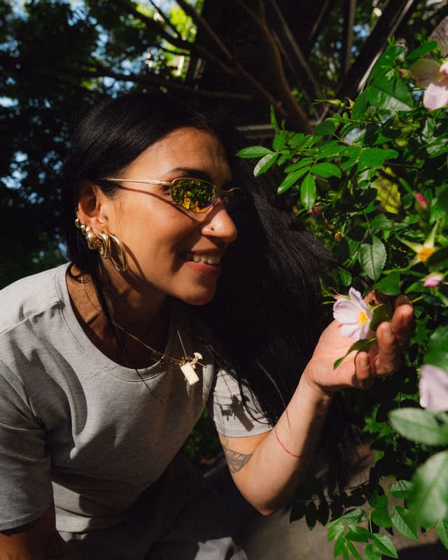 Smiling woman in sunglasses gently touches a pink flower among green leaves.