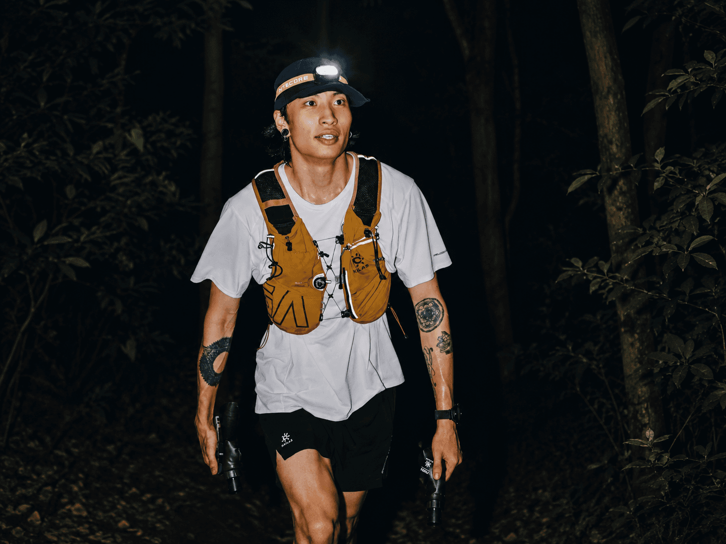 A man trail running at night in a forest, wearing a headlamp and a running vest.