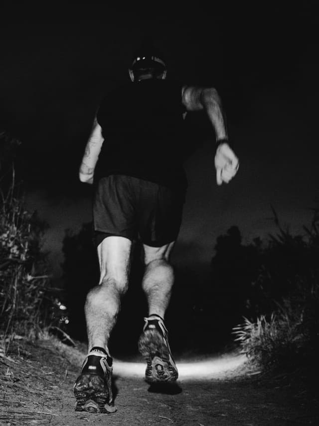 Black and white image of a person running on a dark trail, their headlamp illuminating the path ahead.