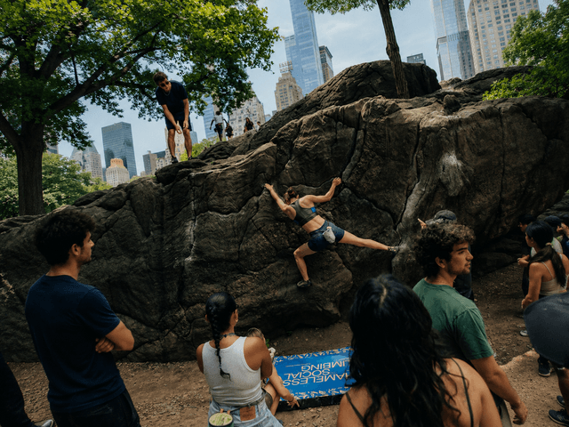 A woman bouldering on a large rock with spectators and city buildings in the background.