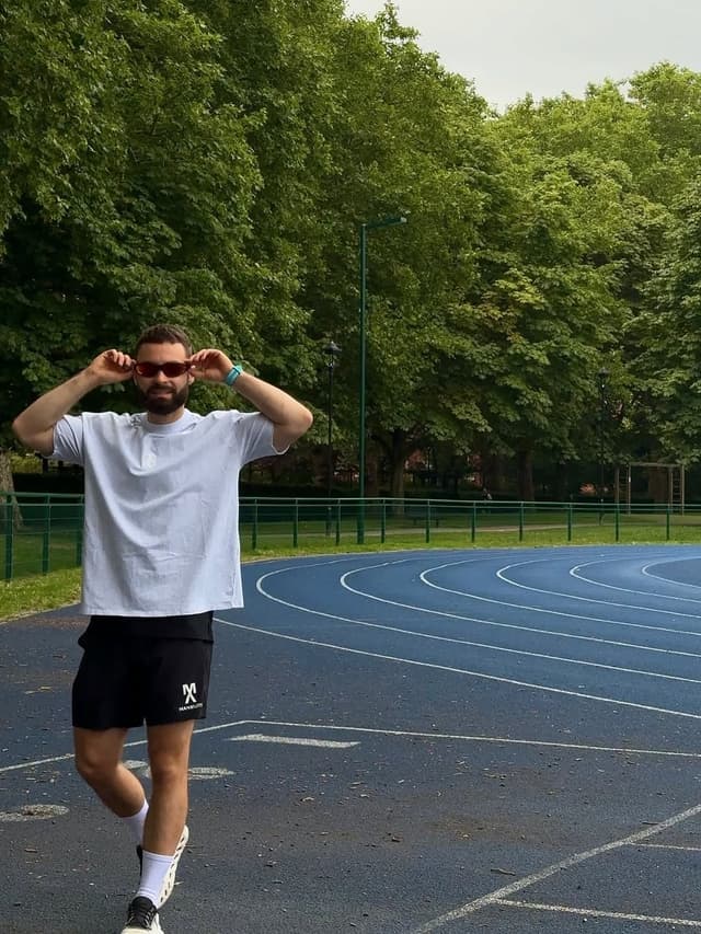 A man in a white t-shirt and black shorts adjusting red sunglasses on a blue running track.