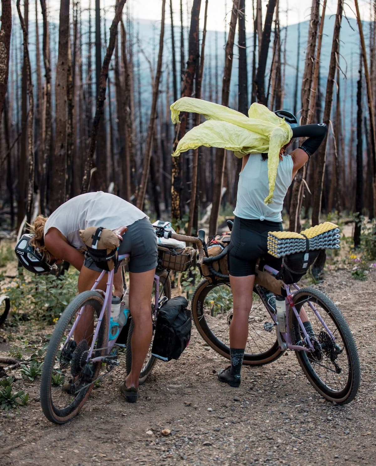 Two bikepackers on a dirt path in a burnt forest. One bends over her bike to adjust gear, while the other puts on a neon yellow jacket.