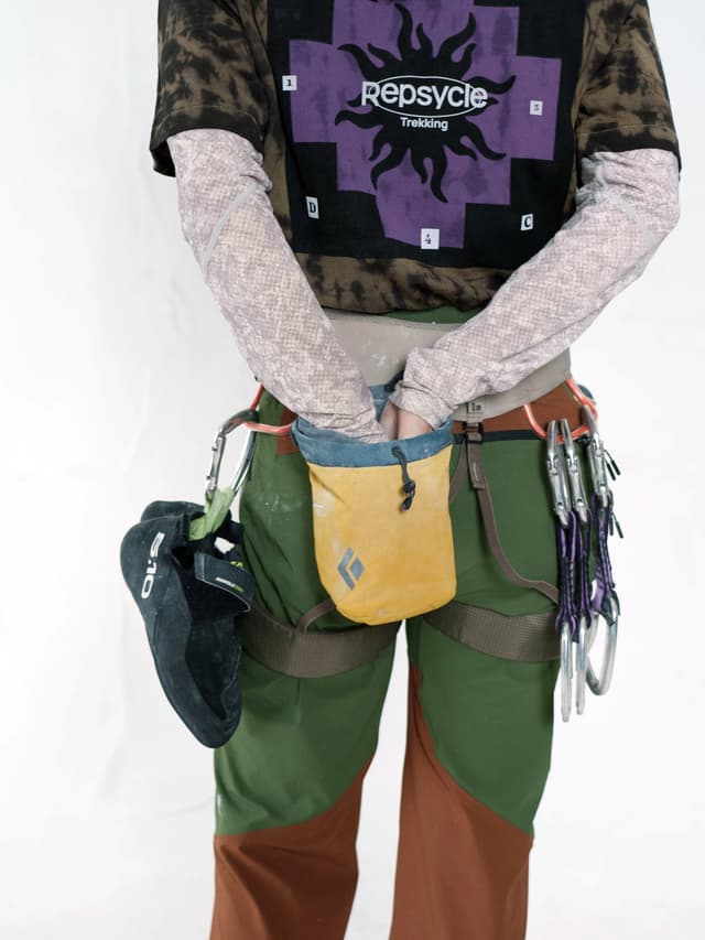 A person wearing climbing gear, with hands in a yellow chalk bag, a black climbing shoe, and carabiners clipped to their harness.
