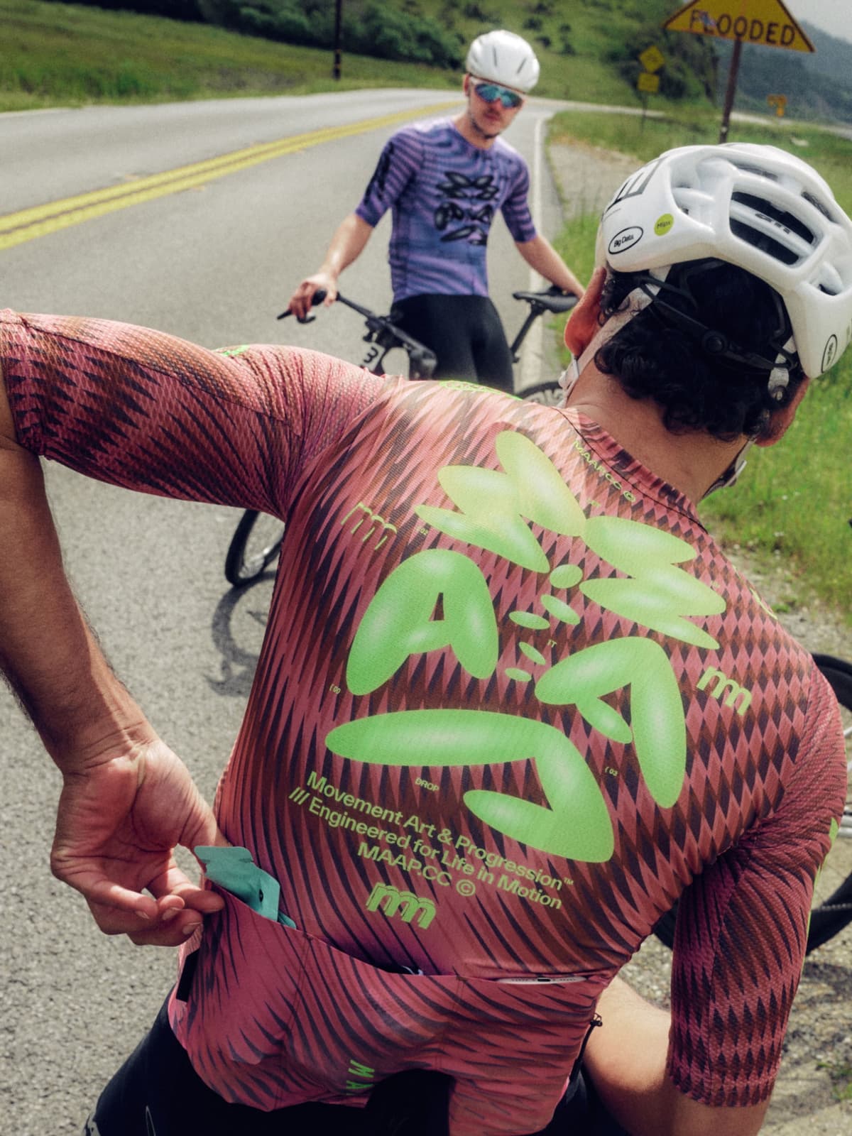 A cyclist in a pink and black patterned jersey with neon green lettering reaches into a back pocket, while another cyclist in a purple jersey stands with their bike on a road with a "FLOODED" sign in the background.