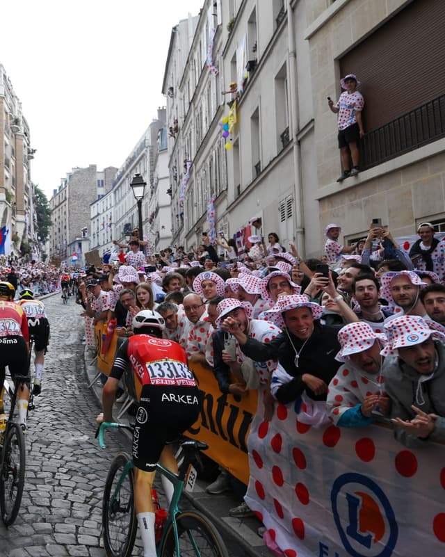 Tour de France cyclists on a cobblestone street lined with cheering fans wearing red polka dot hats and holding matching banners.