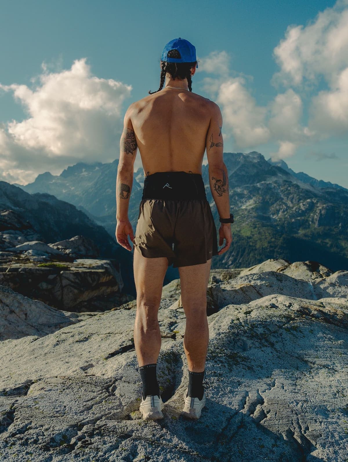 A shirtless person wearing a blue cap and shorts stands on a rocky mountain, looking at a distant mountain range under a partly cloudy sky.