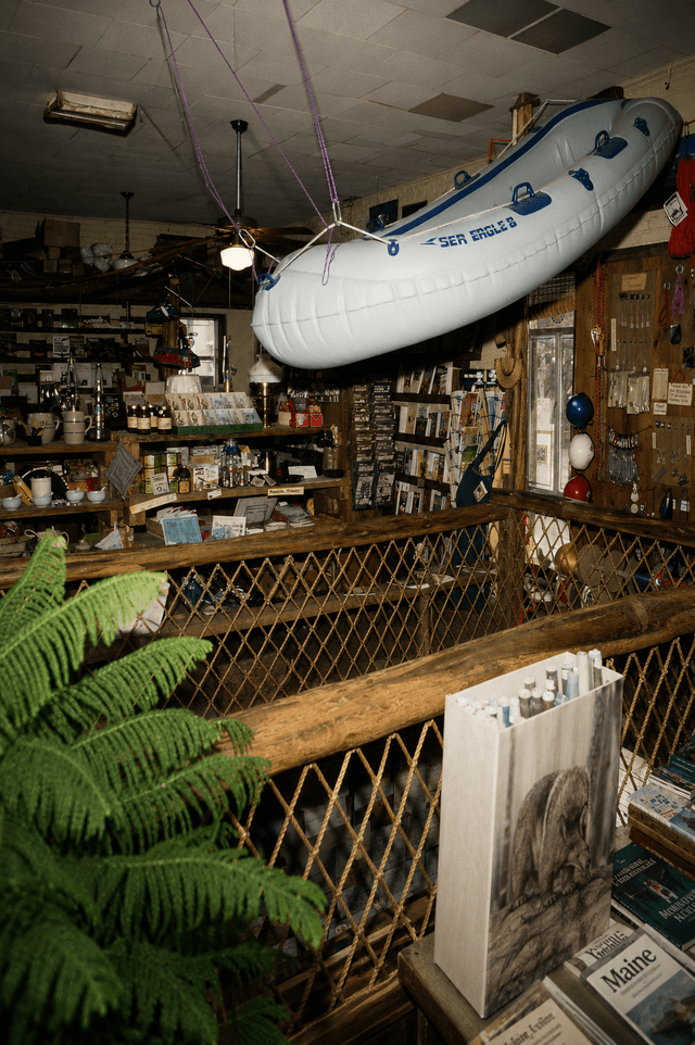 An inflatable raft hangs from the ceiling in a rustic, cluttered store with wooden railings and shelves of various goods.