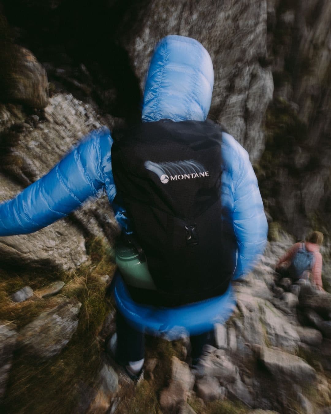 Motion-blurred person in blue puffer jacket and Montane backpack hiking rocky terrain, with another person in the background.