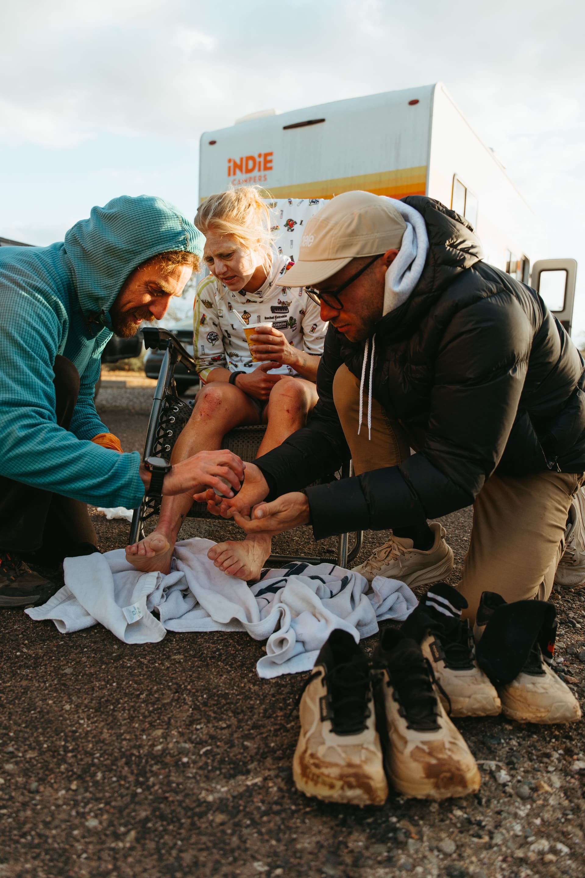 Woman with scraped legs is attended to by two men, with muddy shoes in the foreground and an RV in the background.