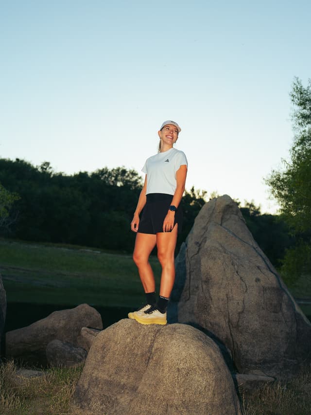 A smiling woman in an Adidas shirt, shorts, and yellow shoes stands on a large rock outdoors.