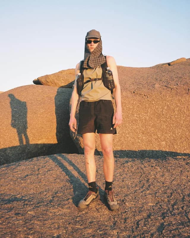 A hiker wearing sunglasses, a plaid sun scarf, and a hydration vest stands on sunlit desert rocks.