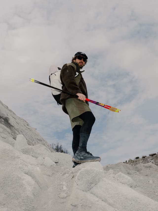 A man in hiking gear with sunglasses and a backpack stands on a pale, rocky terrain, holding colorful trekking poles and looking at the camera.