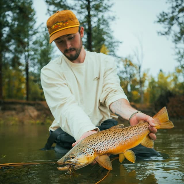 A man in a river holds a brown trout with a lure in its mouth.