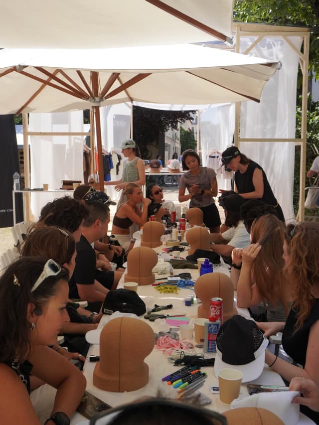 People at an outdoor hat customization workshop, with supplies and head forms on a long table under white umbrellas.