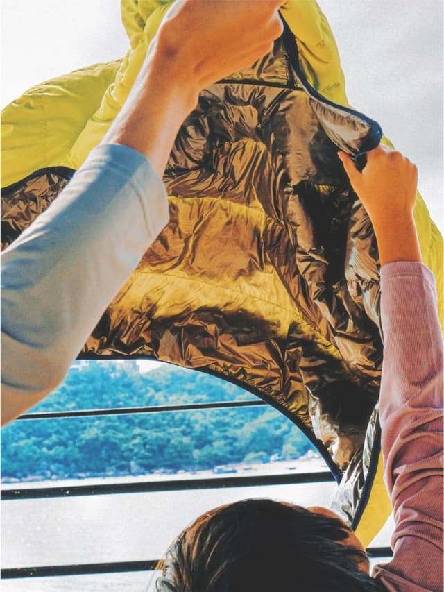 Hands hold open a lime green jacket, revealing its shiny gold lining, with a blurred view of water and green hills below.