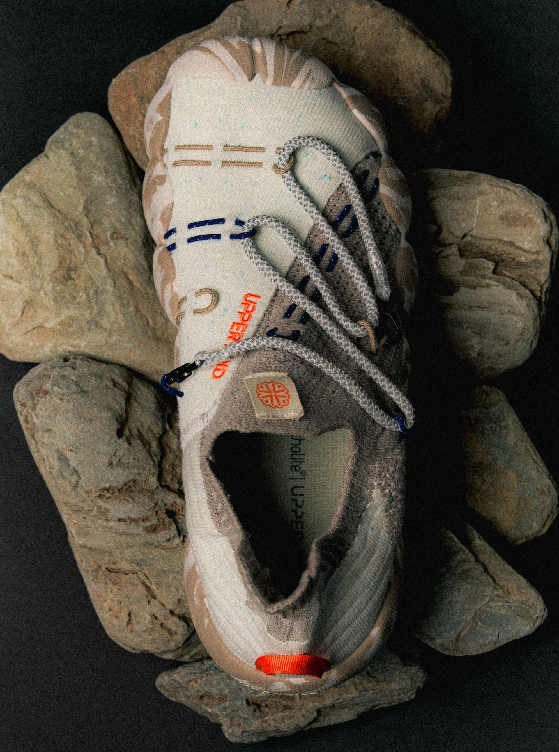Top-down view of a cream and beige knit sneaker with gray laces and an orange pull tab, surrounded by rocks.
