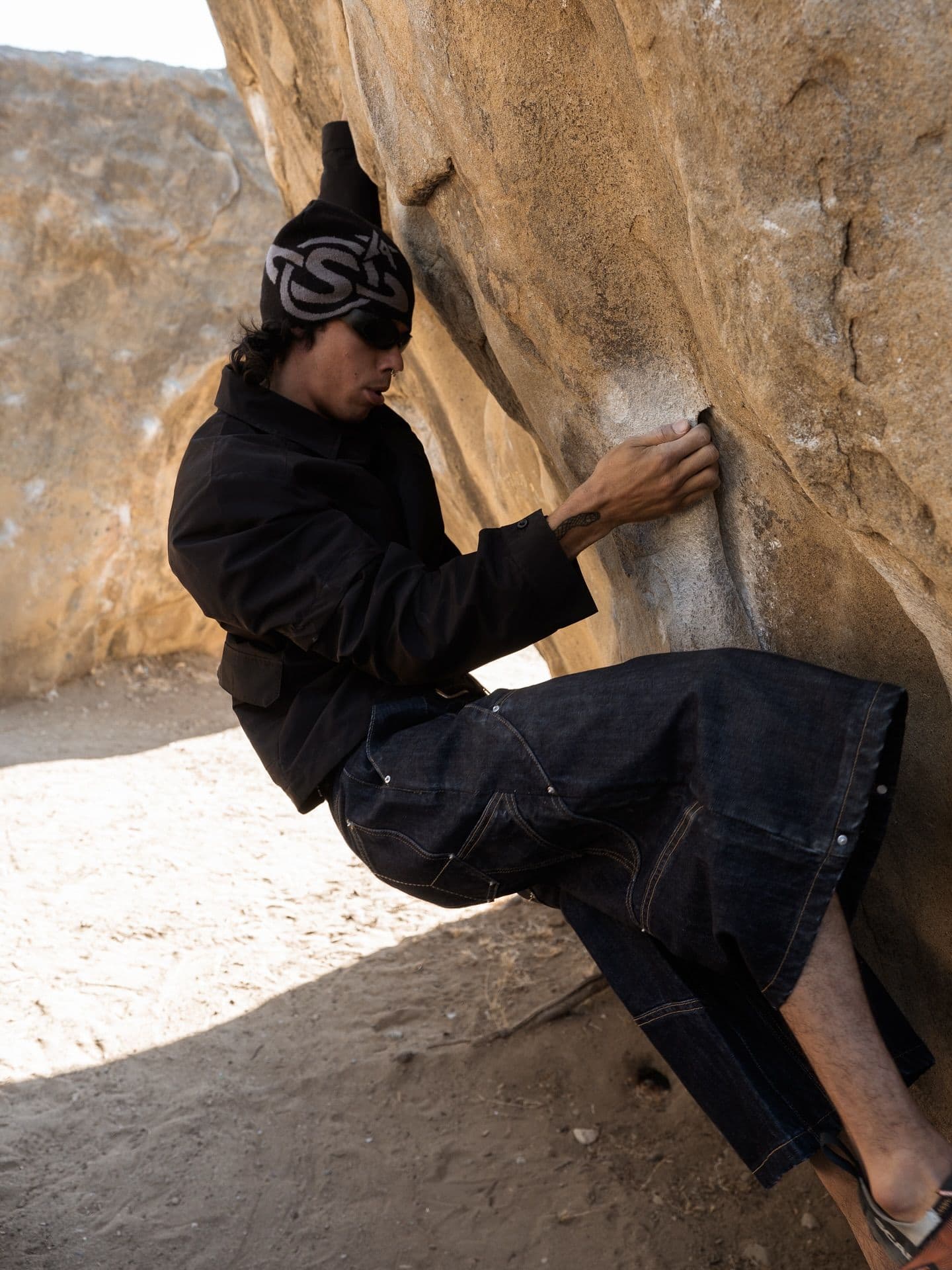 A person in dark clothing and a spiky beanie bouldering on a large light brown rock.