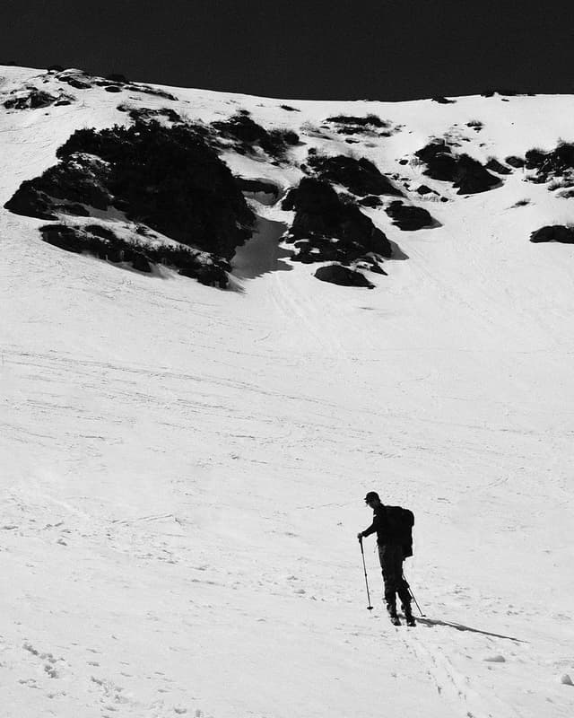 Black and white photo of a ski tourer ascending a snowy mountain slope with dark rock formations under a dark sky.