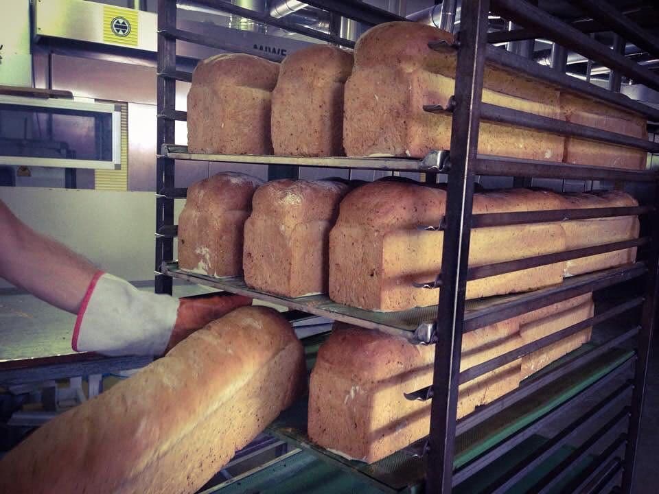 A gloved hand places a loaf of freshly baked bread onto a multi-tiered rack full of other loaves in a bakery.
