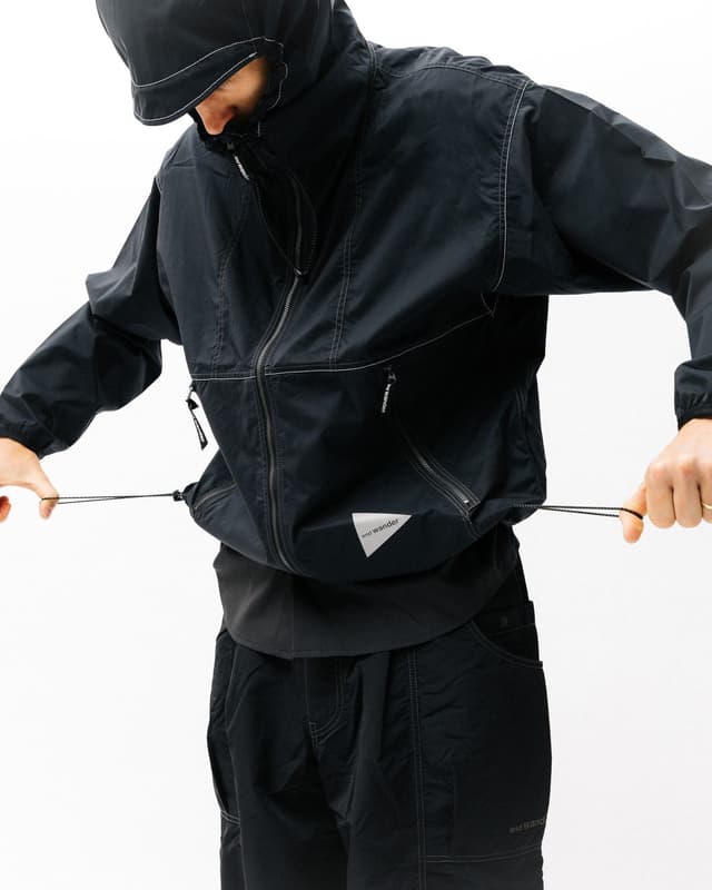 A person in a black hooded jacket with white stitching and an "and wander" logo adjusts the drawstring at the jacket's hem.
